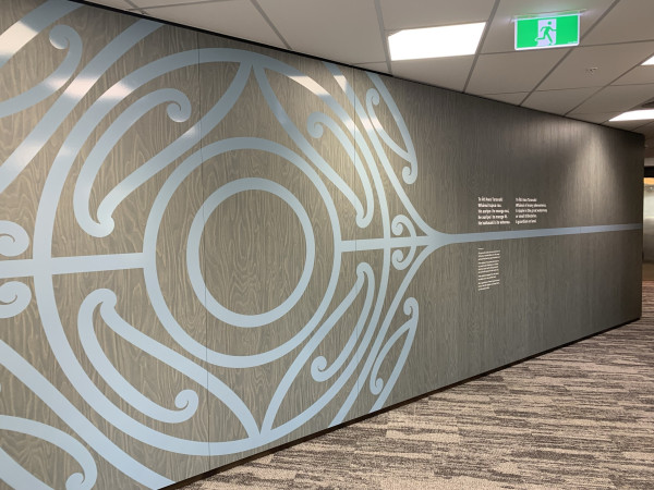 A striking Māori design with circular and spiral patterns in light blue against a grey woodgrain style wall.